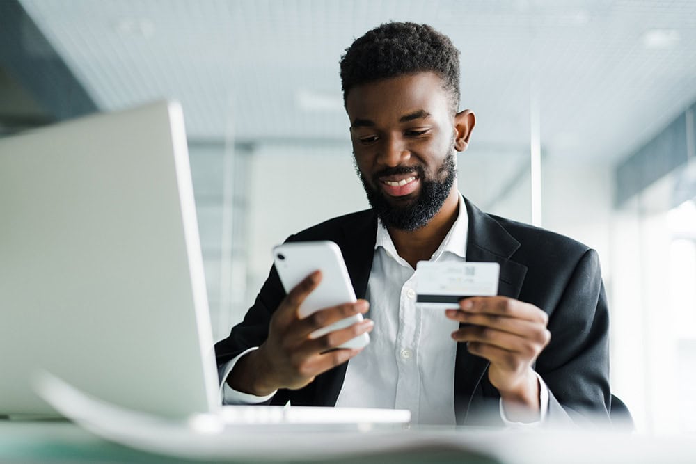 A man sitting in front of his laptop holding his phone in one hand and his credit card in his other hand