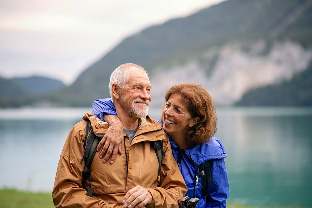 A couple stands in front of a lake and mountain scenery. They are smiling and holding each other