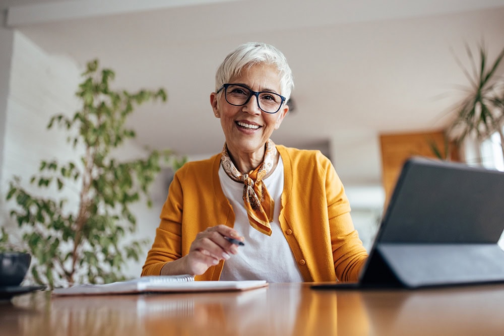 A woman sits at her desk with her iPad to the side and a notebook in front of her. She is smiling