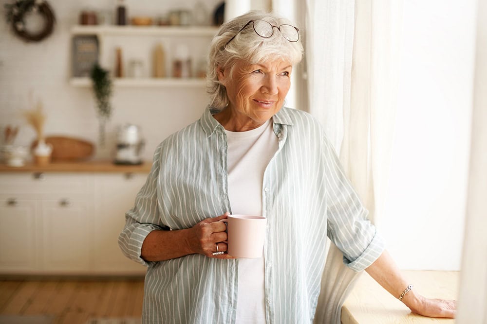 An older woman stands by the window with a cup of coffee in her hand. She is smiling.