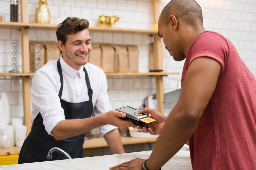 An employee at a coffee shop is handing the card reader to a customer who is paying for his purchase