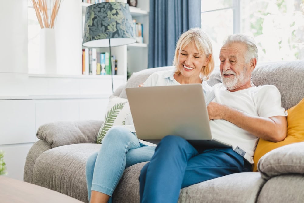 An older couple is sitting on the couch and smiling while looking at their laptop