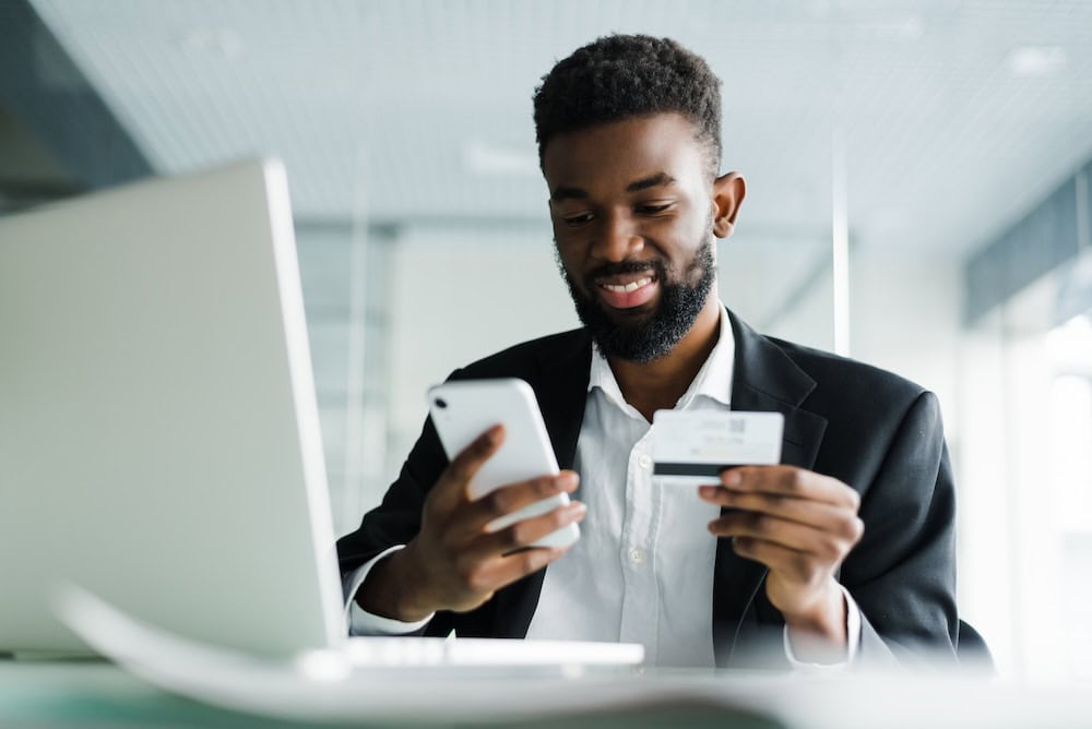 A man is sitting in front of his laptop and smiling while holding his phone in his right hand and his credit card in his left hand