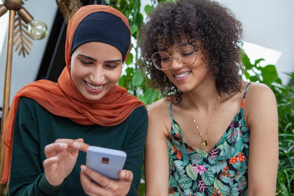 Two women are sitting next to each other and smiling down at the woman on the left's cell phone