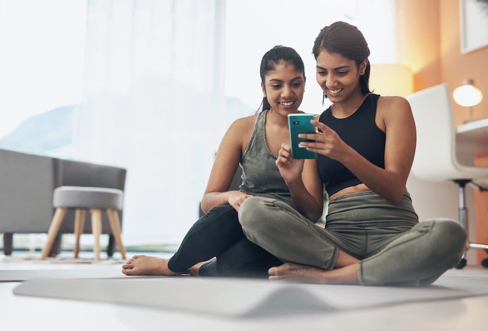 Two women sit on the floor of their home and are smiling while looking at the first woman's cell phone