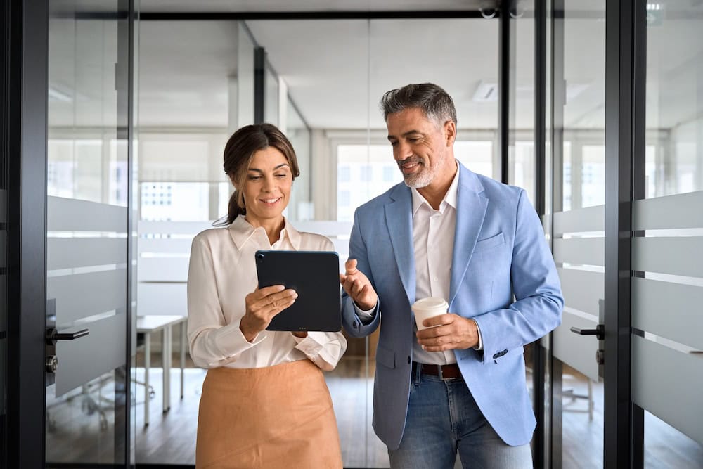 A woman and man are walking through an office while talking and smiling with each other. The woman is holding an iPad and is showing the man something on the screen.